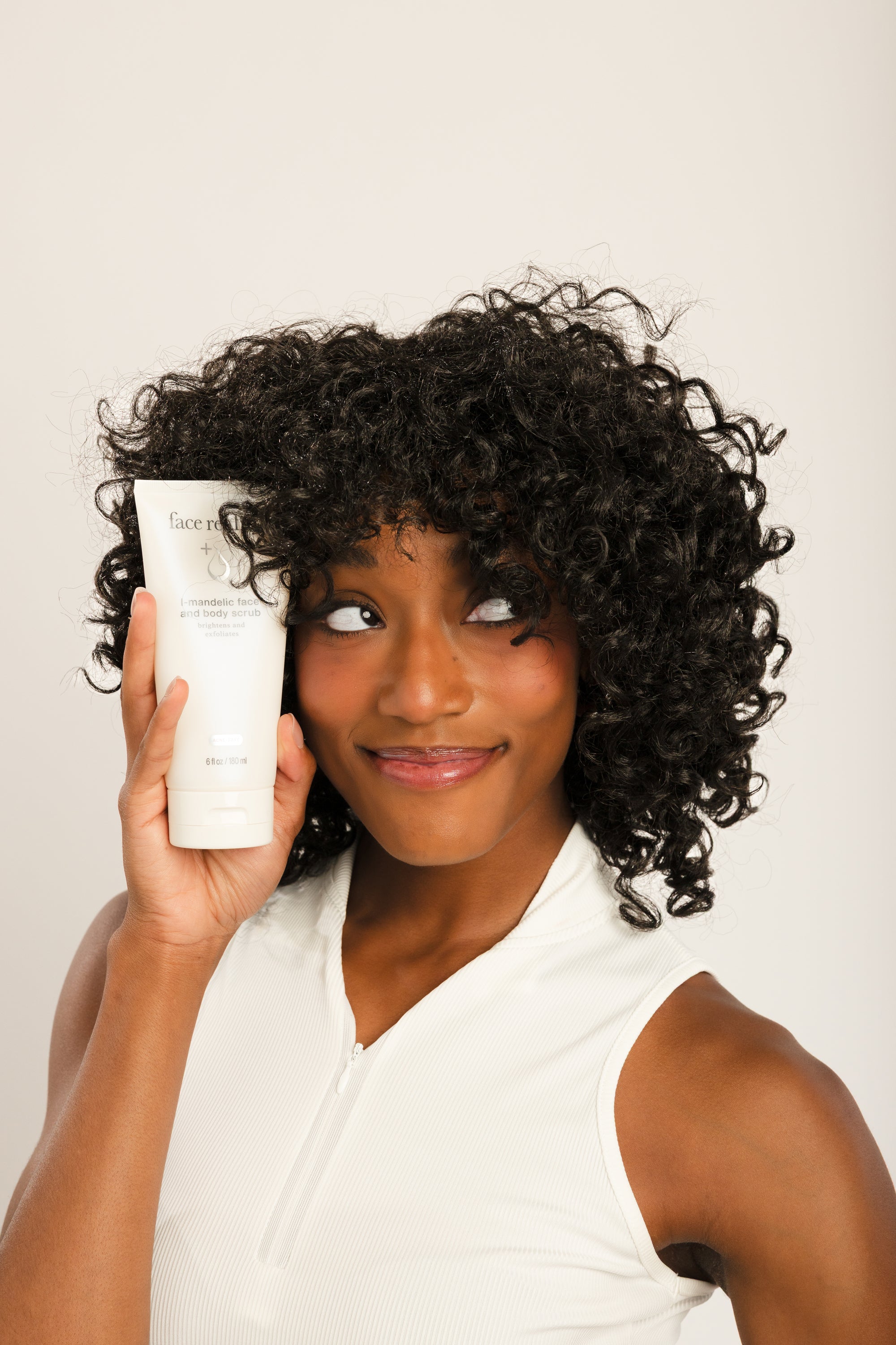 A woman with voluminous dark curly hair holds up a white tube of hair product, looking to the side with a smile against a plain light background.