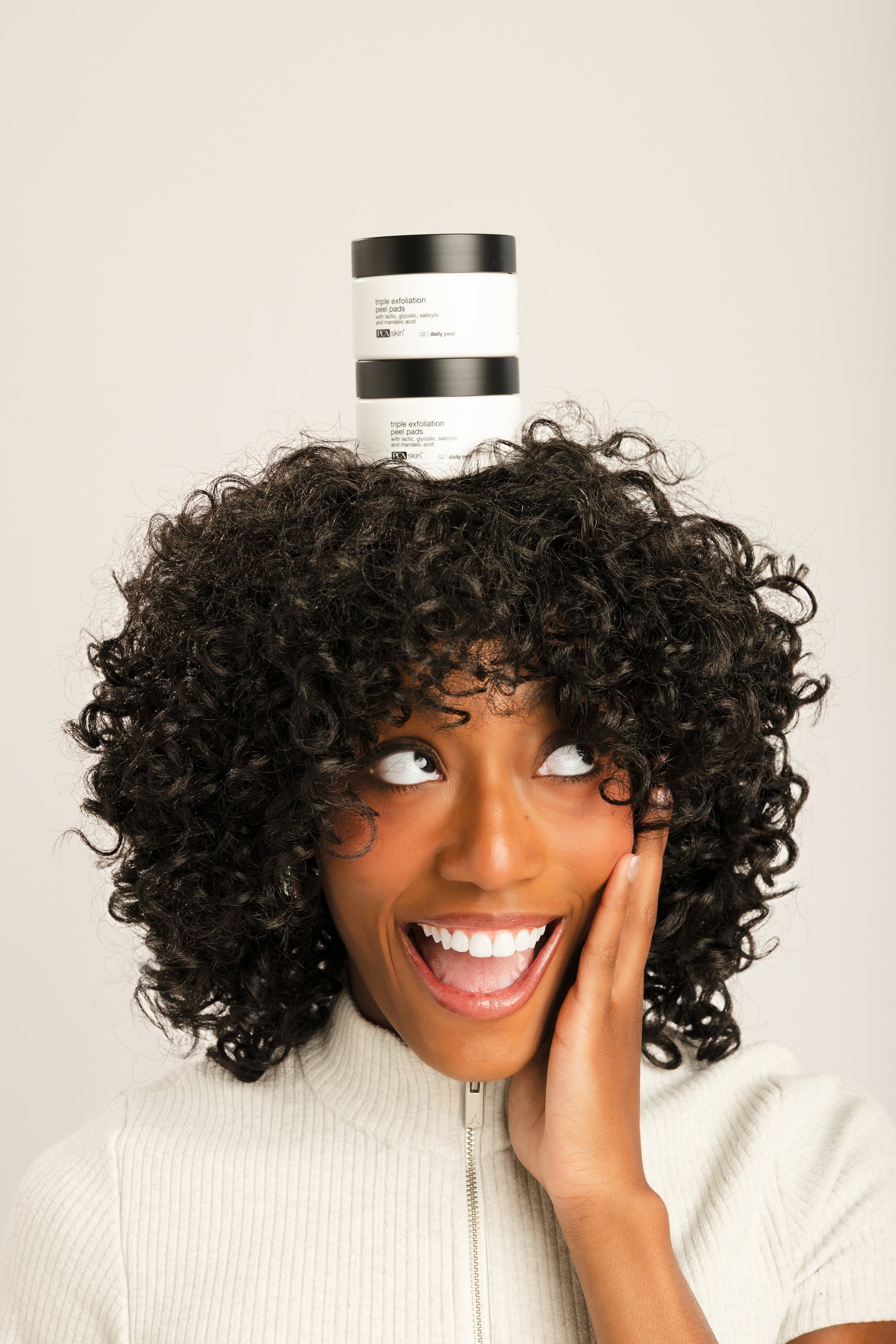 A smiling woman with voluminous dark curly hair balances a stack of two white and black hair product jars on top of her head against a plain light beige studio background.