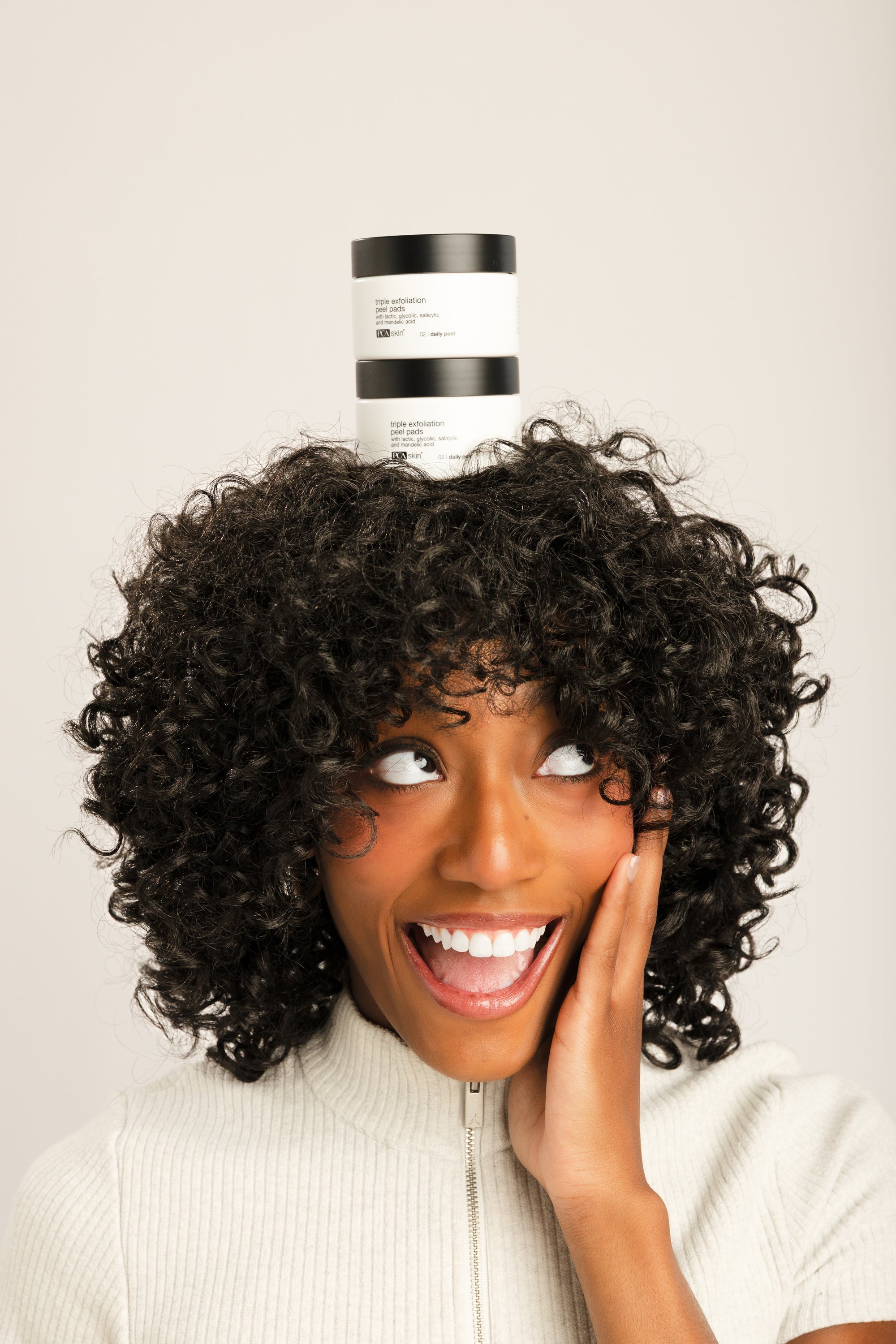 A smiling woman with voluminous dark curly hair balances a stack of two white and black hair product jars on top of her head against a plain light beige studio background.