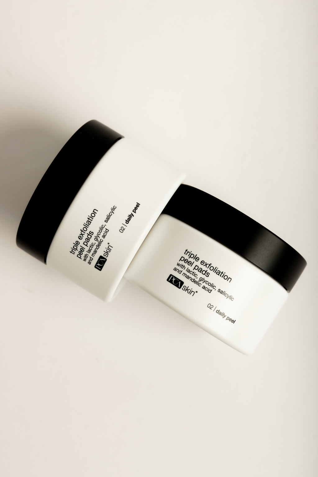 Two stacked white jars of PCA SKIN Triple Exfoliation Peel Pads with black lids and black text, on a cream background